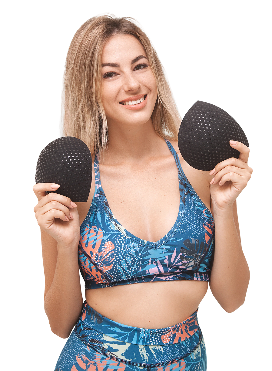 Smiling woman in colorful sports bra and shorts holding black bra inserts, highlighting comfort and grip benefits.
